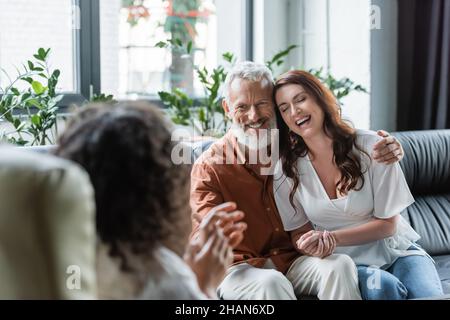 Fröhlicher Mann umarmt glückliche Frau während der Konsultation mit verschwommenen afroamerikanischen Psychologen Stockfoto