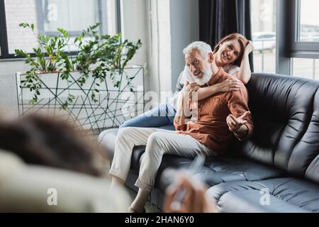 Lächelnde Frau umarmte Ehemann und sprach während der Konsultation mit einem unscharfen afroamerikanischen Psychologen Stockfoto