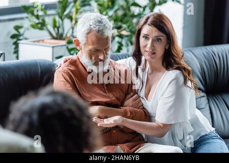 Besorgte Frau, die mit einem verschwommenen Psychologen spricht, während sie einen depressiven Mann umarmt, der mit gekreuzten Armen Stockfoto