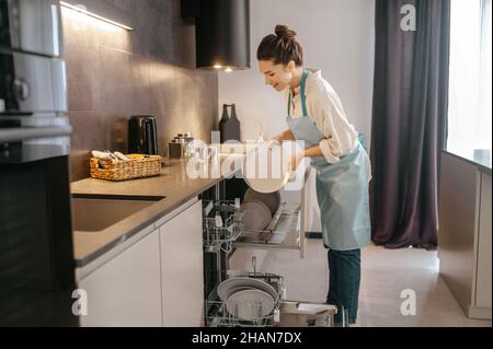 Frau, die in der Nähe der Spülmaschine stand und Teller herausnahm Stockfoto