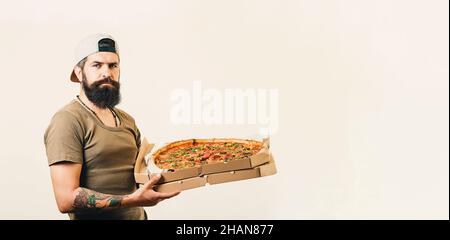 Ein gutaussehender Kurier hält Pappkartons mit Pizza, schlägt vor, leckeres Fast Food zu probieren, trägt Hut und T-Shirt. Stockfoto