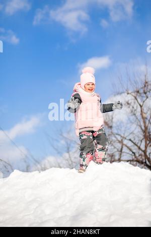 Das kleine Mädchen spielt gegen den blauen Himmel. Kind hat Spaß an sonnigen Wintertag in der Natur, spielt und reitet Schneerutsche, Sprünge auf Schneeverwehungen, Winter Stockfoto