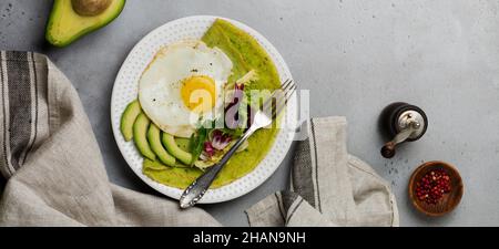 Grüne Pfannkuchen (Spinat) mit Spiegelei, Avocado und Salatblättern auf Keramikplatte auf grauem Betongrund. Konzept der gesunden br Stockfoto