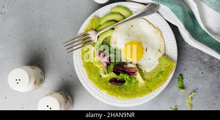 Grüne Pfannkuchen (Spinat) mit Spiegelei, Avocado und Salatblättern auf Keramikplatte auf grauem Betongrund. Konzept der gesunden br Stockfoto