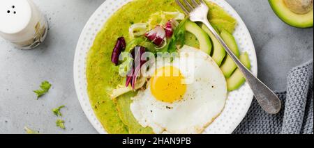 Grüne Pfannkuchen (Spinat) mit Spiegelei, Avocado und Salatblättern auf Keramikplatte auf grauem Betongrund. Konzept der gesunden br Stockfoto