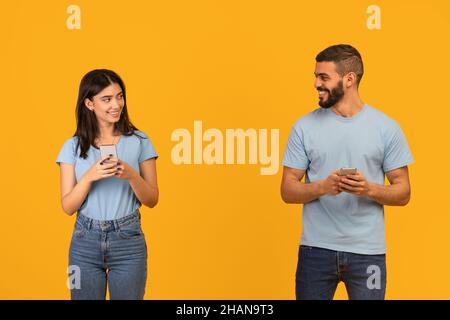 Soziale Distanz und moderne Online-Kommunikation. Glückliche arabische Ehegatten tippen auf dem Telefon, schauen und lächeln sich an Stockfoto