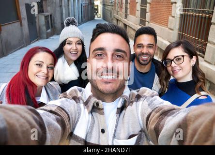 Positive multiethnische Freunde, die nahe stehen und weit lächeln, während sie in einer engen Straße Selbstporträts machen und die Kamera betrachten Stockfoto