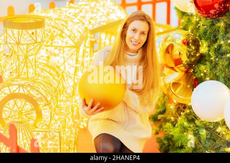 Lächelnde Dame, die einen riesigen mattierten Goldball hält, während sie neben einem Weihnachtsbaum und einem funkelnden dekorativen Zug sitzt. Feieratmosphäre. Herzlichen Glückwunsch Stockfoto