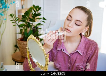 Schöne junge Frau macht erröten auf ihrem Gesicht mit Make-up Pinsel, während zu Hause in einem Raum an einem Tisch sitzen. Kaukasisches Mädchen in einem dunkelrosa Hemd macht Make-up auf eigene Faust. Stockfoto