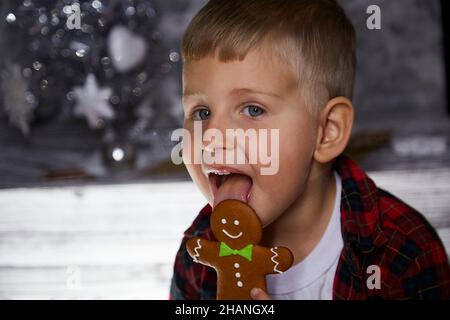 Nettes glückliches Kind leckt Lebkuchen und macht einen lustigen Gesichtsausdruck in der Nähe von Weihnachtsbaum. Stimmungsvolles, ästhetisches Zuhause für Silvester. Fröhlicher Bab Stockfoto