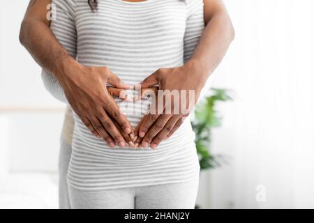 Der kurz geschnittene junge afroamerikanische Ehemann umarmt seine Frau mit herzförmigen Händen auf dem Bauch in einem minimalistischen Schlafzimmer Stockfoto