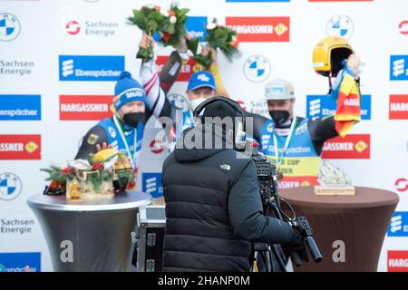 Altenberg, Deutschland. 12th Dez 2021. Rennrodel: WM, Einzel, Herren, zweite Runde. Ein Kameramann steht während der Preisverleihung vor Max Langenhan (l-r) aus Deutschland, Wolfgang Kindl aus Österreich und Johannes Ludwig aus Deutschland. Quelle: Sebastian Kahnert/dpa-Zentralbild/ZB/dpa/Alamy Live News Stockfoto