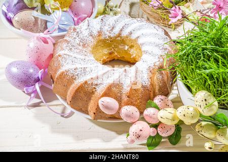 Traditionelle Frühlingsferien Osterrunde Kuchen mit Puderzucker gekrönt. Auf hellem Holzhintergrund mit Sonnenlicht, Ostereiern, Dekor und Frühlingszwirnholz Stockfoto