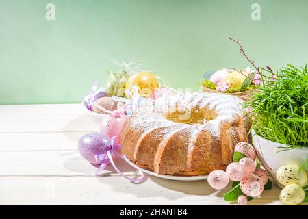 Traditionelle Frühlingsferien Osterrunde Kuchen mit Puderzucker gekrönt. Auf hellem Holzhintergrund mit Sonnenlicht, Ostereiern, Dekor und Frühlingszwirnholz Stockfoto