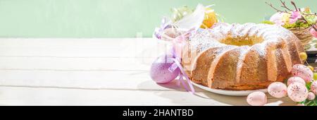 Traditionelle Frühlingsferien Osterrunde Kuchen mit Puderzucker gekrönt. Auf hellem Holzhintergrund mit Sonnenlicht, Ostereiern, Dekor und Frühlingszwirnholz Stockfoto