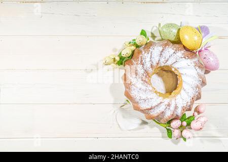 Traditionelle Frühlingsferien Osterrunde Kuchen mit Puderzucker gekrönt. Auf hellem Holzhintergrund mit Sonnenlicht, Ostereiern, Dekor und Frühlingszwirnholz Stockfoto