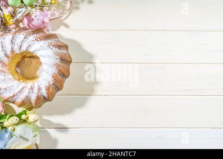 Traditionelle Frühlingsferien Osterrunde Kuchen mit Puderzucker gekrönt. Auf hellem Holzhintergrund mit Sonnenlicht, Ostereiern, Dekor und Frühlingszwirnholz Stockfoto