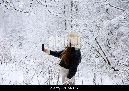 Mädchen in schwarzem Mantel und Pelzhut Selfie auf Smartphone-Kamera auf dem Hintergrund der Schnee Natur im Winterwald. Bäume nach Schneefall Stockfoto