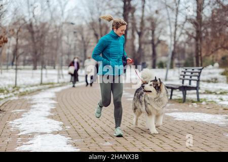 Die Sportlerin joggt zusammen mit ihrem Hund im Park bei schneebedecktem Wetter. Winterfitness, Haustiere, Freundschaft Stockfoto