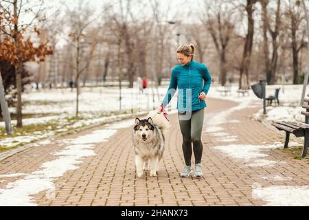 Die Sportlerin joggt zusammen mit ihrem Hund im Park bei schneebedecktem Wetter. Winterfitness, Haustiere, Freundschaft Stockfoto