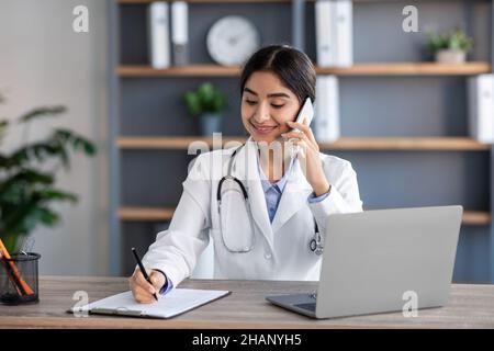 Lächelnd hübsche junge hindu-Ärztin im weißen Mantel mit Stethoskop telefoniert mit dem Patienten, arbeitet im Büro Stockfoto