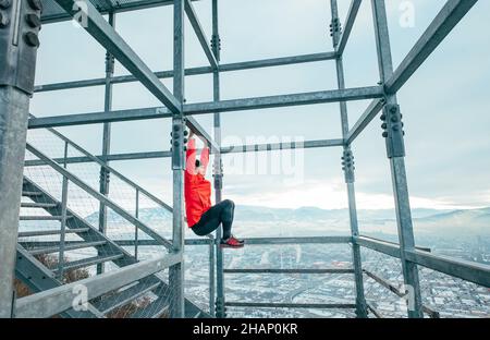 Ein frecher Mann, der in leuchtend roter, sportlicher Kleidung und Laufschuhen gekleidet ist, die durch Anheben der Beine eine horizontale Stange zum Training der bauchmuskeln aufhängen. Fitnesstraining auf der Stahlschale Stockfoto