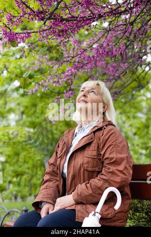 Die fröhliche blonde Frau aus dem Jahr 50s mit einem transparenten Regenschirm sitzt auf einer Holzbank in einem städtischen Sommerpark neben einem blühenden Baum Stockfoto