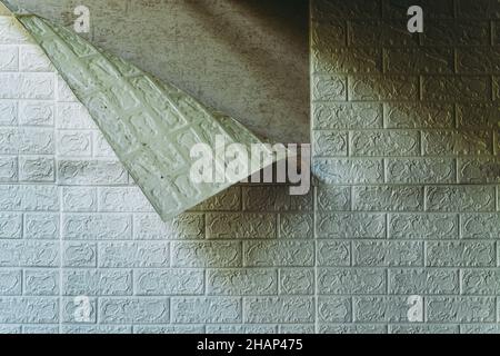 Die Tapete beginnt sich abzulösen. Die Tapete aus alten weißen Ziegeln schälte sich von der Wand im Raum, weil es feucht war oder sich nicht gut klebte. Reparieren Sie A Stockfoto
