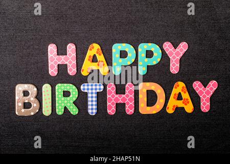 Karte mit Happy Birthday-Wörtern aus gemischten, lebendigen farbigen Holzbuchstaben auf einem strukturierten, dunkelschwarzen Textilmaterial, das als Botschaft verwendet werden kann Stockfoto
