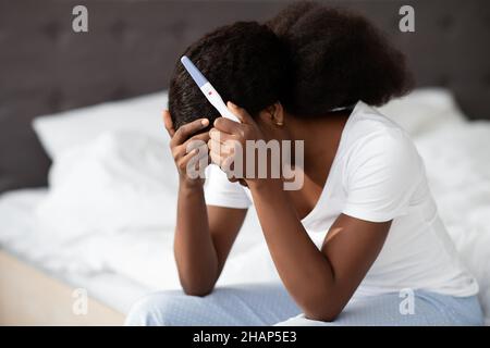 Verzweifelte afroamerikanische Dame, die mit einem Schwangerschaftstest auf dem Bett sitzt Stockfoto