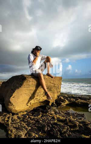Junger Mann mit Hipster- oder Hippie-Auftritt, mit beiden Händen, die sein Gesicht besorgt auf einem Felsen am Strand sitzen Stockfoto