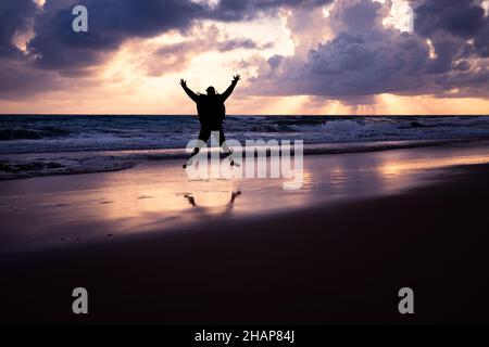 Silhouette eines jungen Mannes, der bei Sonnenaufgang mit ausgestreckten Armen und Beinen an die Küste springt. Stockfoto