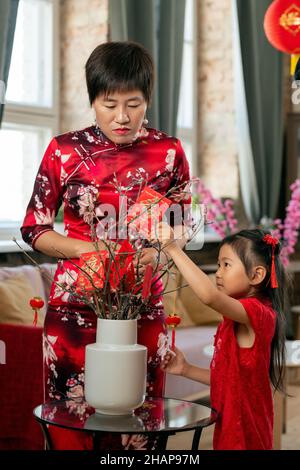 Junge asiatische Frau, die ihre kleine Tochter ansieht, die Postkarten mit Neujahrgrüßen auf Äste legt Stockfoto