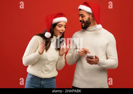 Glücklicher arabischer Mann, der eine Nachricht erhält, die dem überraschten Weib ein Smartphone zeigt, das Santa-Hüte trägt, roter Studiohintergrund Stockfoto