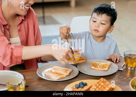 Glückliche asiatische Frau hilft ihrem kleinen Sohn, Sandwich mit Honig oder Marmelade beim Frühstück zu machen Stockfoto