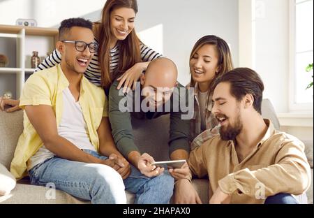 Lächelnder Mann hält Smartphone und zeigt seinen fröhlichen Freunden lustige Fotos während der Party zu Hause. Stockfoto