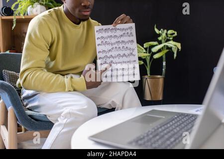 Junge selbstbewusste Lehrerin, die während des Online-Unterrichts im Sessel vor dem Laptop auf Notenblätter zeigt Stockfoto