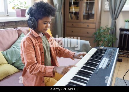 Glücklicher biracial fleißiger Junge, der die Tasten der Klaviertastatur drückt, während er auf der Couch im großen Wohnzimmer sitzt Stockfoto