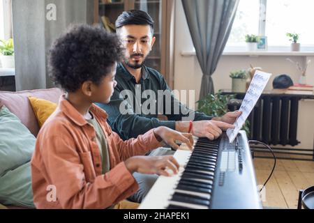 Junge Musiklehrerin, die auf ein Notenblatt zeigt, während sie auf einen niedlichen fleißigen Schuljungen schaut, der Klaviertastatur spielt Stockfoto