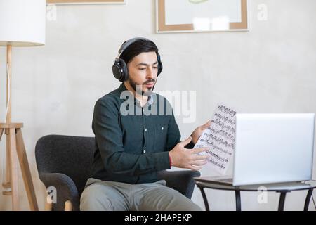 Junge Musiklehrerin in Kopfhörern, die Noten mit Noten zeigt, während sie mit dem Online-Publikum kommuniziert Stockfoto