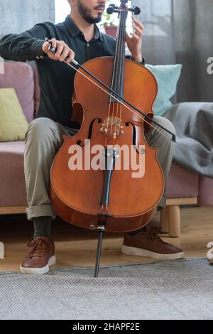 Junger Mann in Casualwear sitzt auf der Couch in häuslicher Umgebung und spielt Streichinstrument Stockfoto