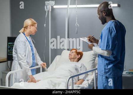 Reife Ärztin im Gespräch mit einer älteren Frau über die Operation, während afrikanische Anästhesistin Notizen in der Krankenkarte macht Stockfoto