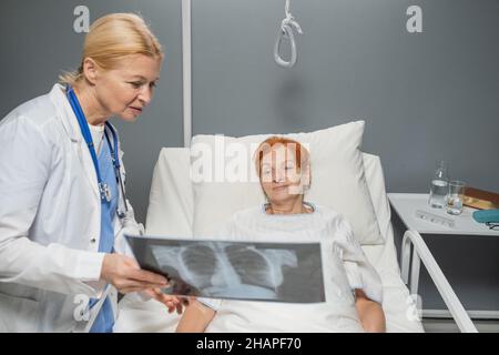 Reifer Arzt im weißen Laborkittel, der das Röntgenbild einer älteren Frau zeigt, die auf der Krankenhausstation im Bett liegt Stockfoto