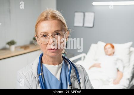 Porträt einer reifen Ärztin in einer Brille, die auf die Kamera schaut, während sie auf der Krankenstation steht und die Patientin im Hintergrund hat Stockfoto