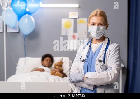 Porträt eines reifen Arztes in Schutzmaske, der die Kamera mit gekreuzten Armen auf der Krankenstation betrachtet und den Patienten im Hintergrund sieht Stockfoto