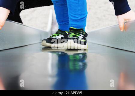 Teilansicht eines Kindes auf einer Spielplatzrutsche, Spiegelung in Metalloberfläche. Stockfoto