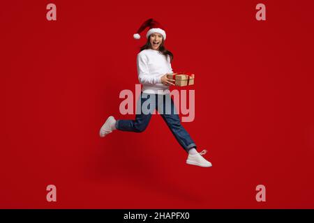 Positive Millennial-Frau Mit Weihnachtsmütze, Die In Der Luft Springt, Mit Geschenkbox Stockfoto