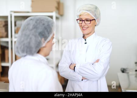 Blonde kaukasische Angestellte in steriler Uniform, die mit dem Vorgesetzten über die Qualität der Produkte spricht und die gekreuzten Arme hält. Innenraum der Lebensmittelfabrik. Stockfoto