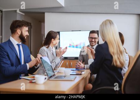 Lächelnder CEO sitzt mit seinem Team im Sitzungssaal. Das Team klatschte ihm zu. Ein Meeting ist ein Ereignis, bei dem die Protokolle aufbewahrt werden und die Stunden verloren gehen. Stockfoto