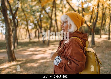 Blonde 50s-Hündin in stilvoller Kleidung mit modischem Lederrucksack geht im Herbst im gordes Park spazieren. Sonnenstrahlen durch die Äste von Bäumen. Reisen genießen Stockfoto